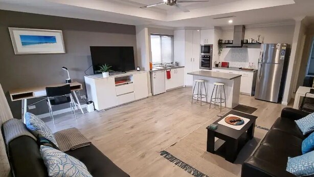 A modern living room with a sofa, a coffee table, and a TV. There is a kitchen area with a fridge, microwave, and sink. - Self Contained Granny Flat with Play Space (Alkimos)