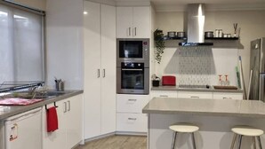 A modern kitchen with white cabinets, a stainless steel refrigerator, a microwave, and an oven. There are two stools at the counter. - Self Contained Granny Flat with Play Space (Alkimos)