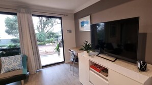 A living room with a large flat-screen TV, a sofa, a coffee table, and a sliding glass door leading to an outdoor area. - Self Contained Granny Flat with Play Space (Alkimos)