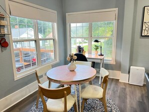 A dining area with a round table, chairs, a vase with flowers, and a view through large windows.