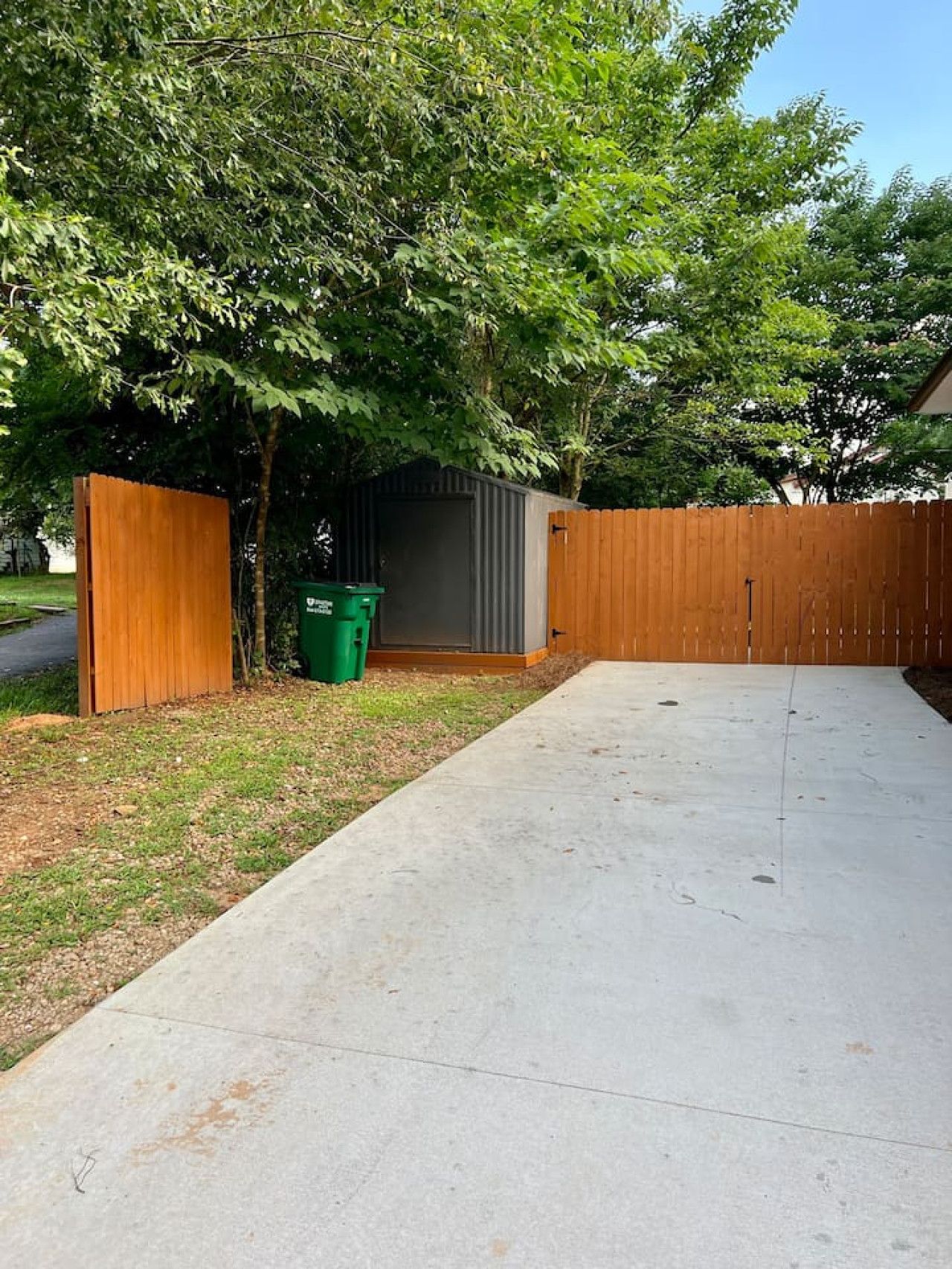 A concrete driveway leading to a wooden fence, a green trash bin, and a grey shed.