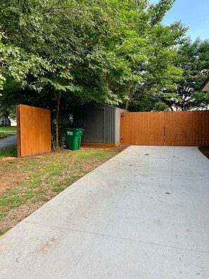 A concrete driveway leading to a wooden fence, a green trash bin, and a grey shed.