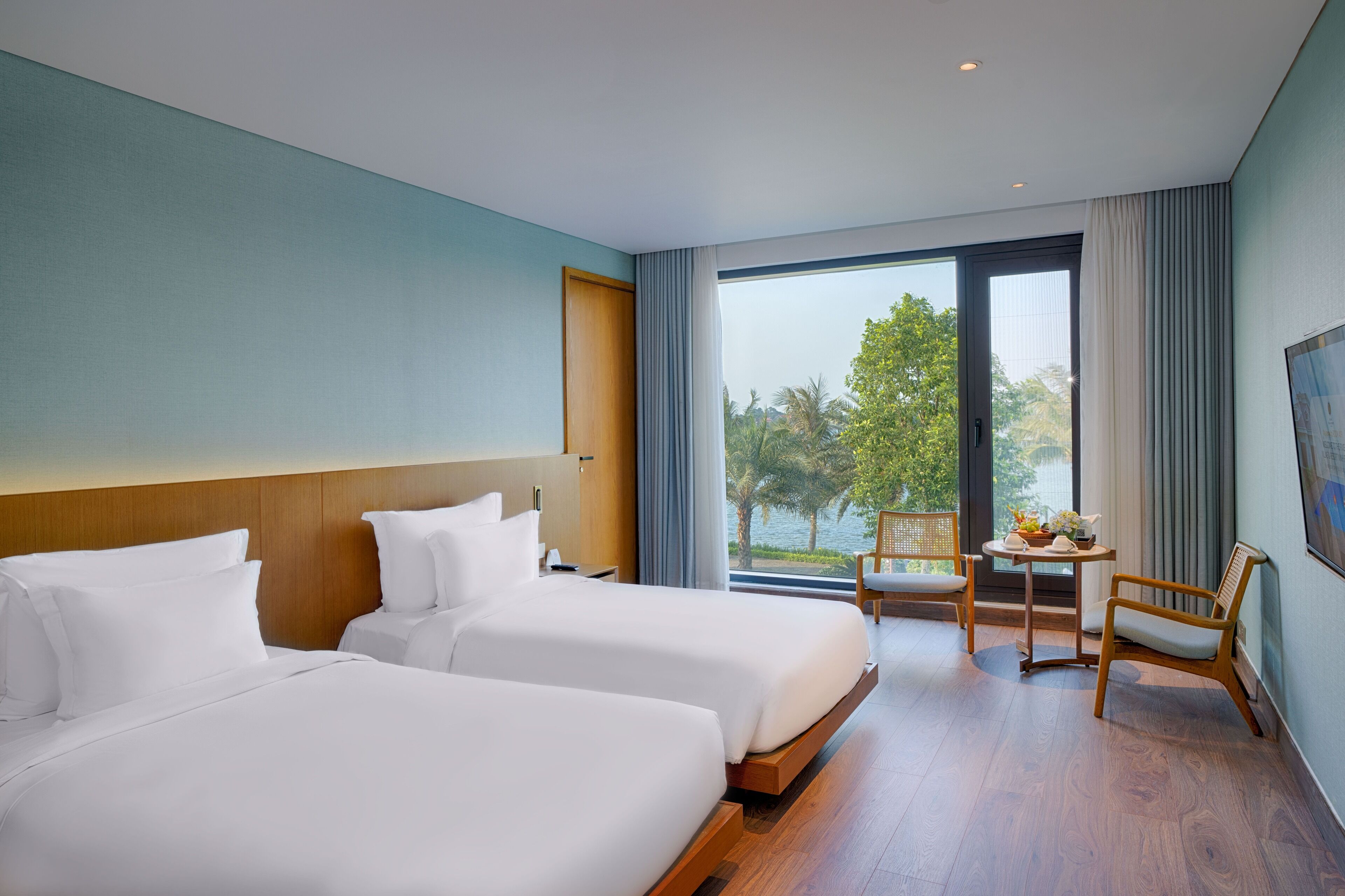 Une chambre d’hôtel avec deux lits, une petite table avec des chaises et une grande fenêtre donnant sur un paysage de verdure et d’eau.