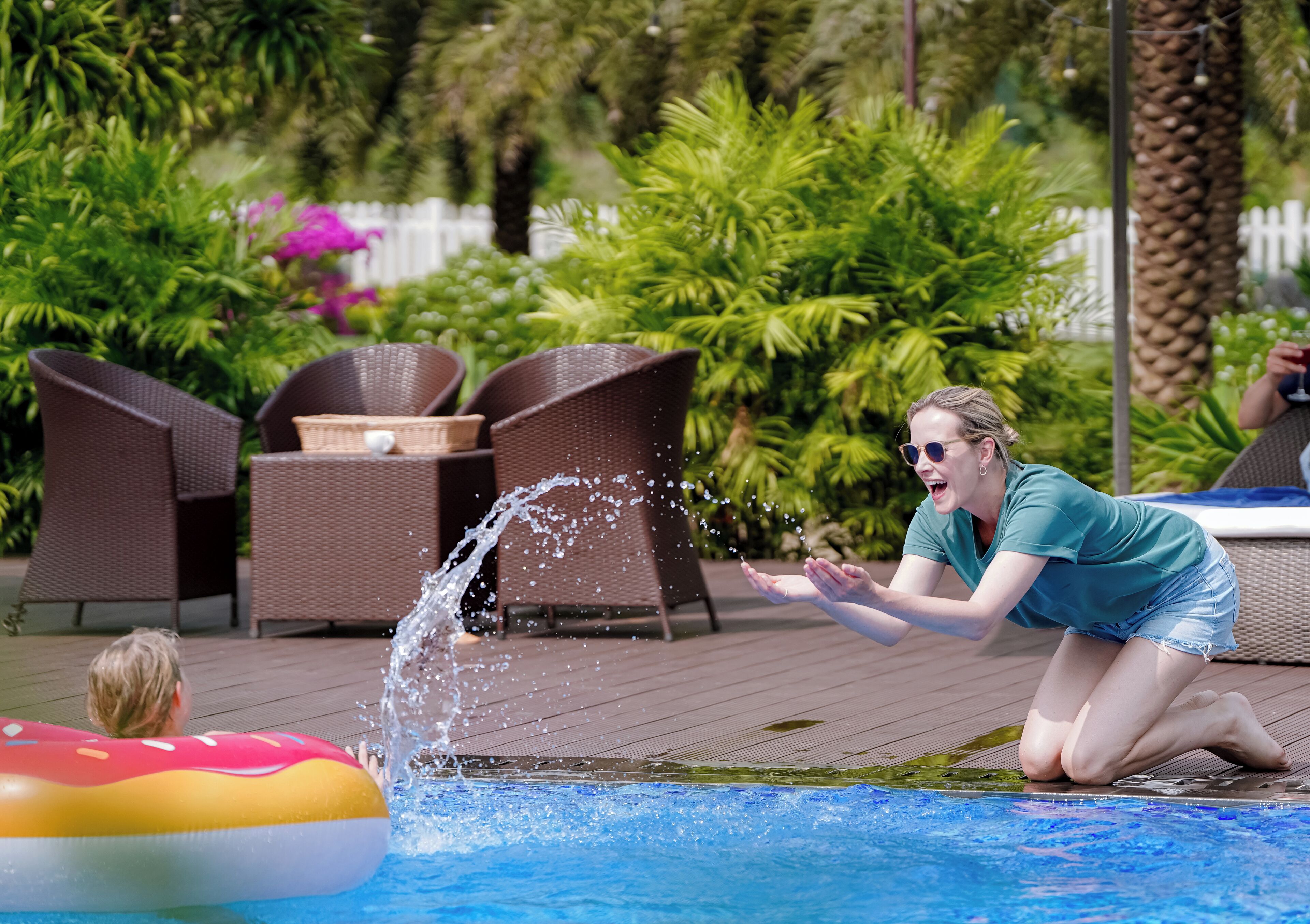 A pool area with a person splashing water, lounge chairs, and a white fence in the background.