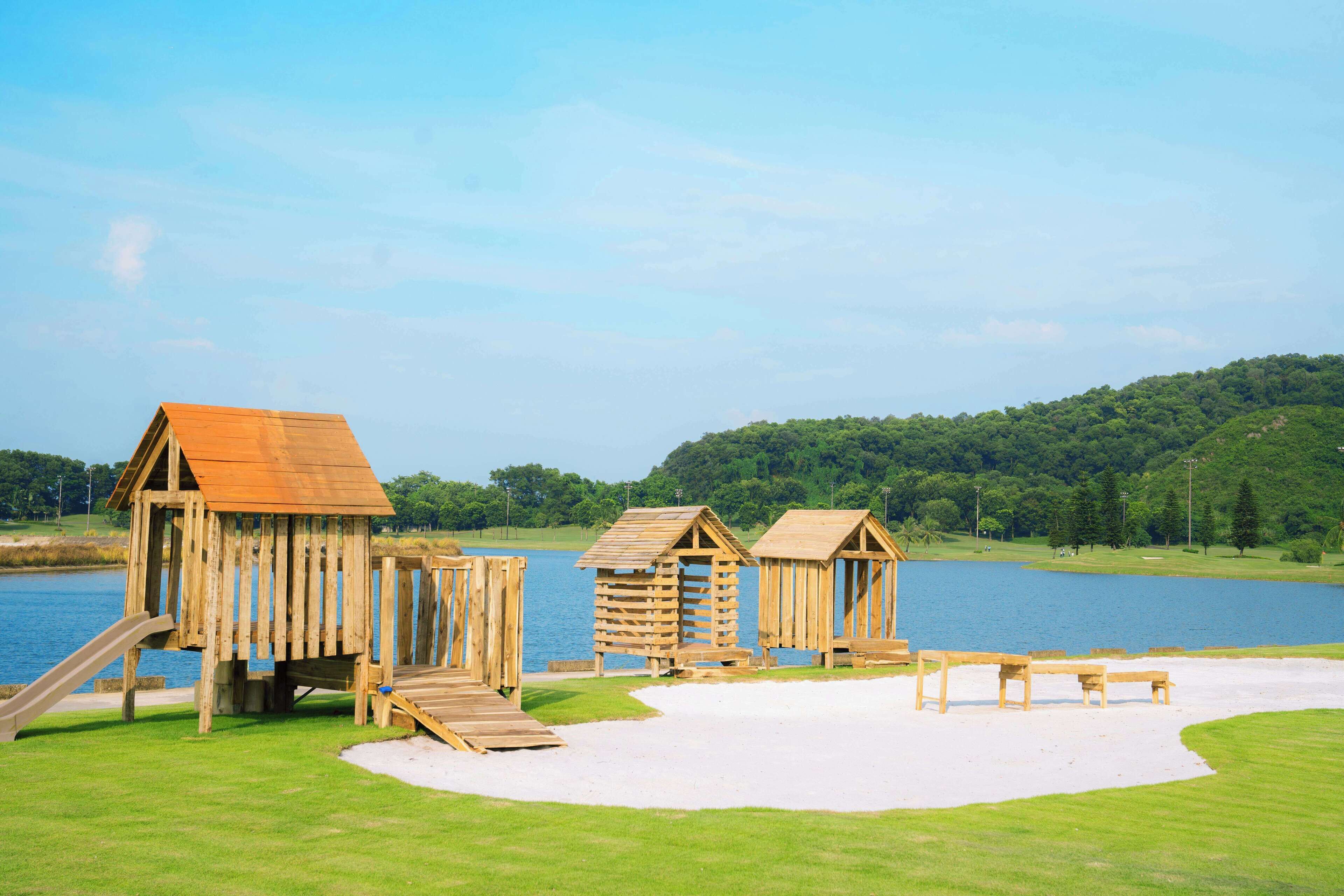 A wooden play structure with a slide and a small bridge, situated near a lake with a grassy area and benches.