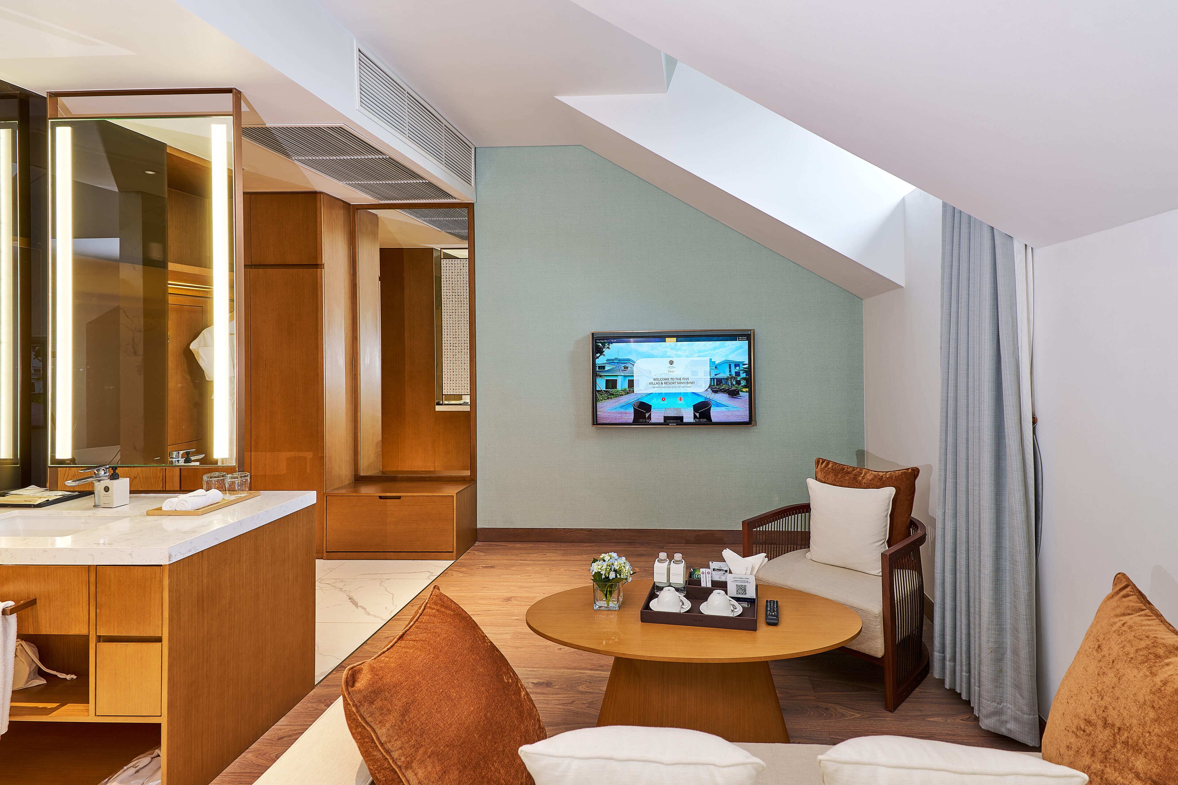 A modern hotel room with a large mirror, a television on the wall, a round wooden table with a tray of toiletries, and a seating area with two chairs.