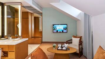 A modern hotel room with a large mirror, a television on the wall, a round wooden table with a tray of toiletries, and a seating area with two chairs.