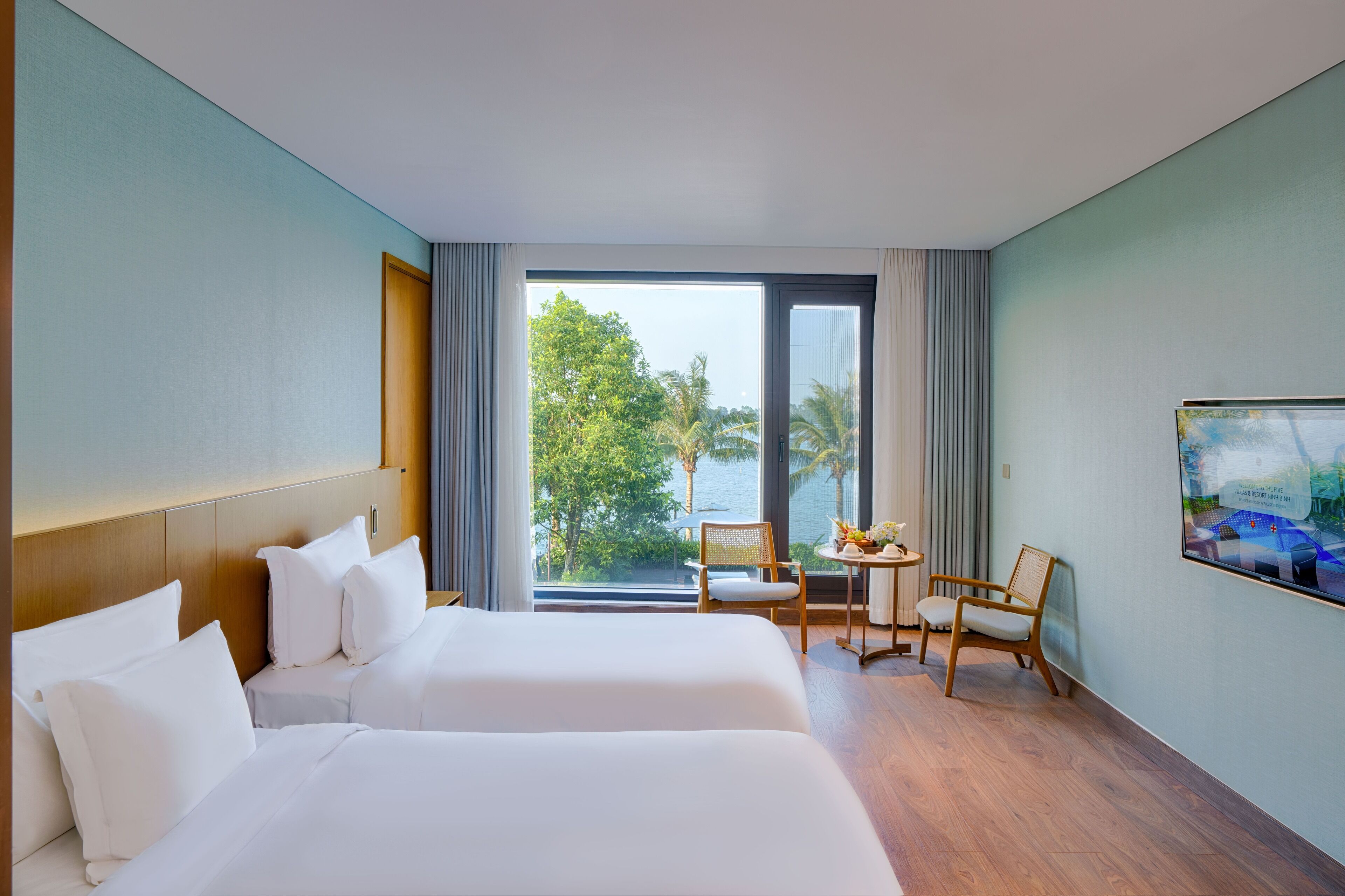 A hotel room with two beds, a small table with chairs, a TV, and a large window with a view of palm trees and water.