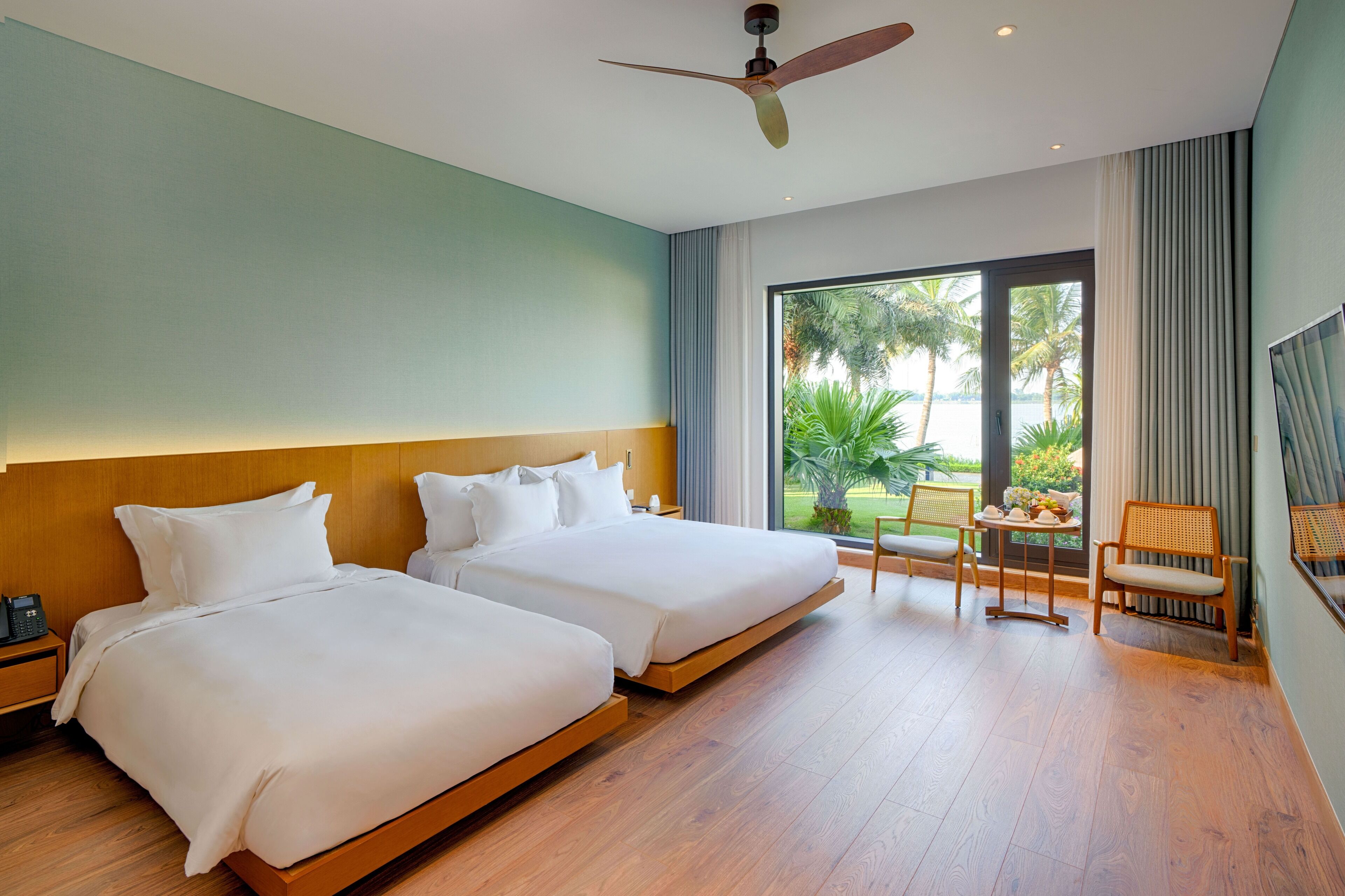 Habitación de hotel con dos camas, un ventilador de techo, un ventanal con vistas a palmeras y una pequeña mesa con sillas.