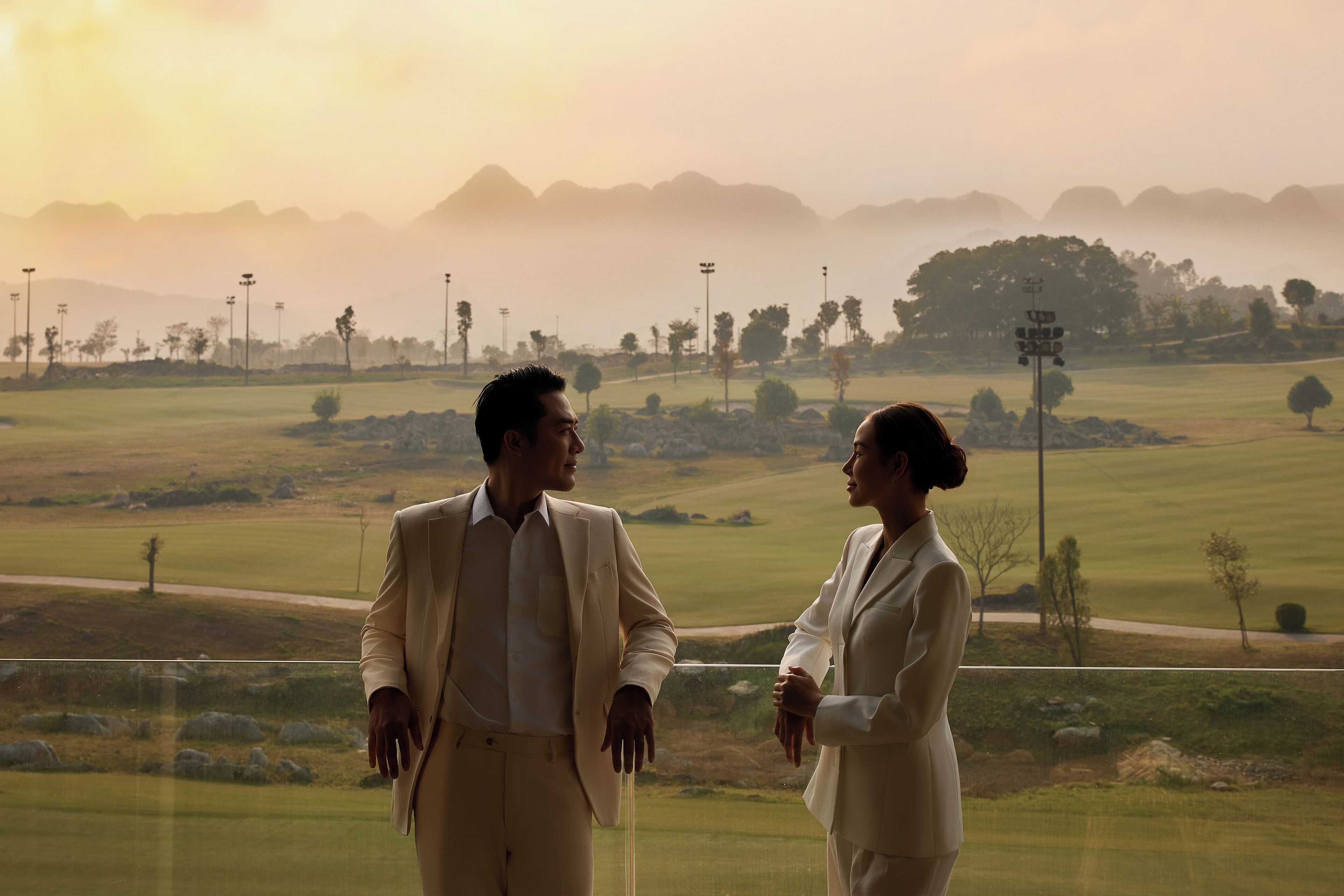 A couple standing on a balcony overlooking a golf course with distant mountains.