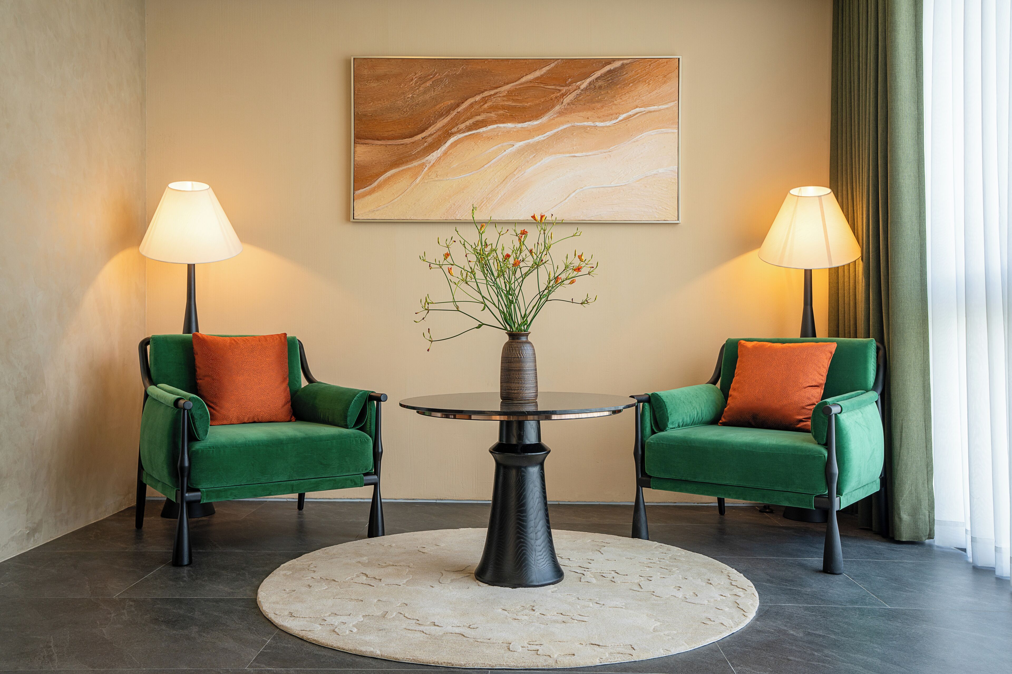 A room with two green armchairs, a round table with a vase of flowers, and two lamps.
