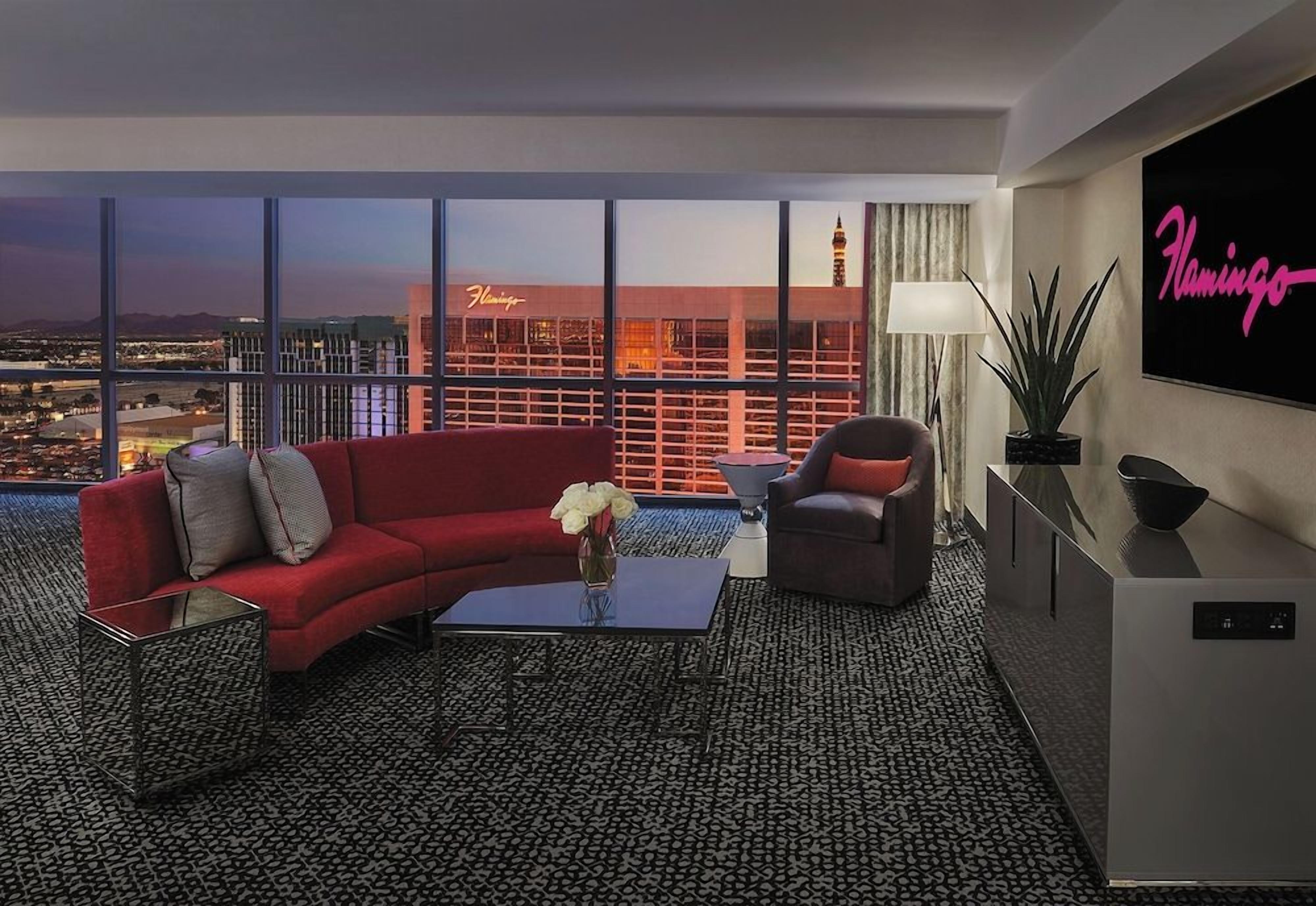 A modern hotel room with a city view, a red sofa, a coffee table, a TV, and a plant.