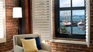 Quarto com vista para um porto, apresentando uma parede de tijolos, uma janela com persianas, uma cadeira com almofadas e um abajur.