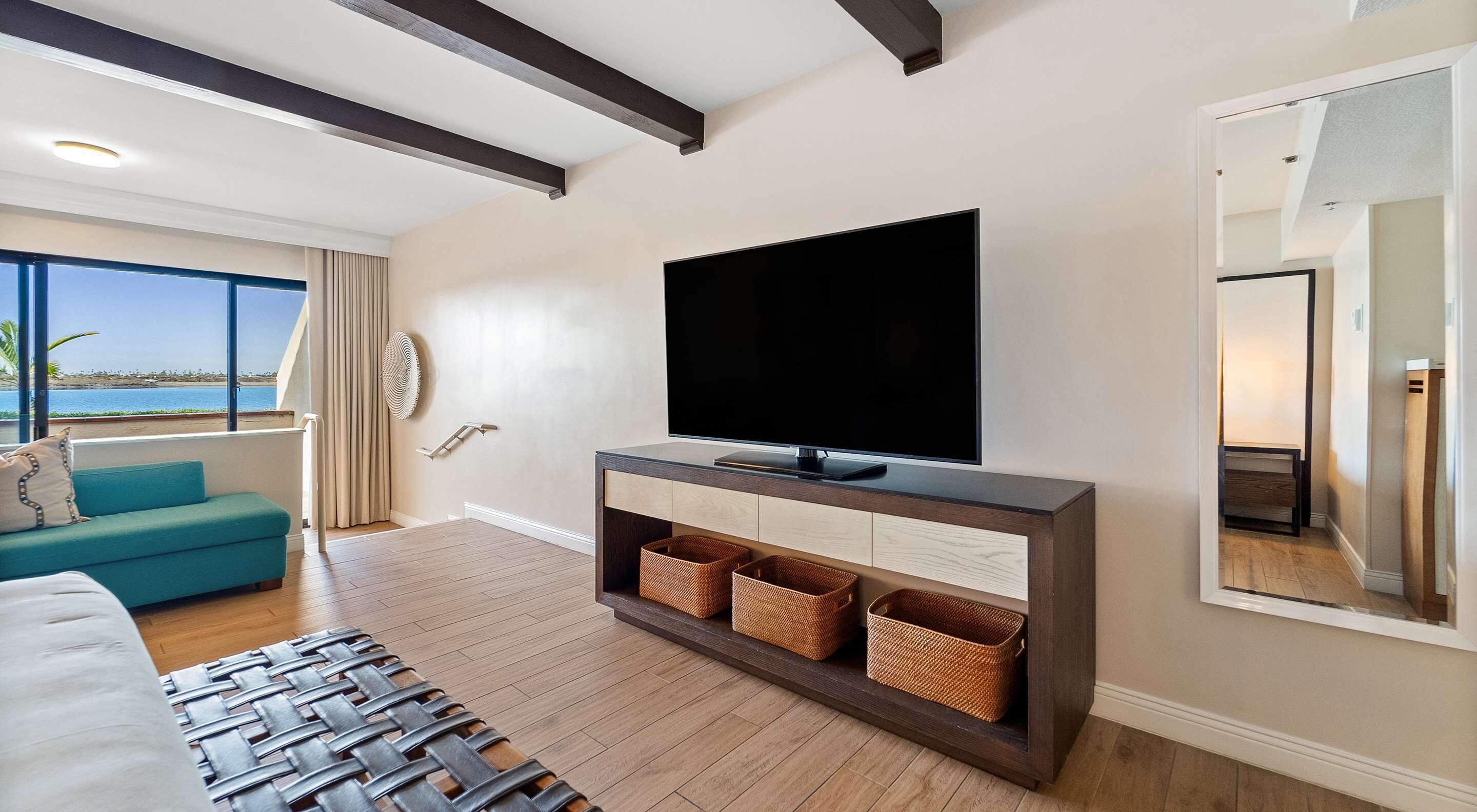 Ein modernes Wohnzimmer mit einem großen Flachbildfernseher, Holzböden, einem türkisfarbenen Sofa und Meerblick durch große Fenster.