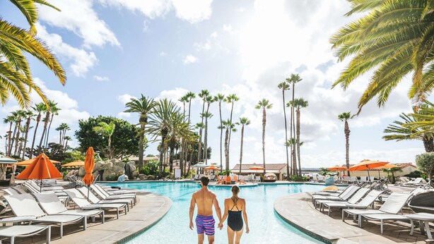 A couple walking towards a pool with lounge chairs and umbrellas around it.