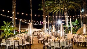 An outdoor dining area with tables set for guests, surrounded by palm trees and string lights.