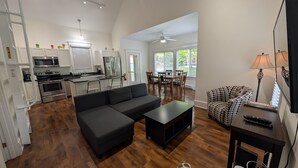 A modern living room with a sectional sofa, a dining area with wooden chairs, and a kitchen with white cabinets and stainless steel appliances.