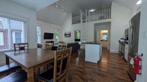 A spacious living area with a dining table, chairs, a sofa, and a TV. There is a kitchen area with a refrigerator and a fire extinguisher.