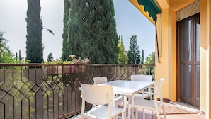 A balcony with a white table and chairs, a metal fence, and tall trees in the background. - Relais Vittoriale (Gardone Riviera)