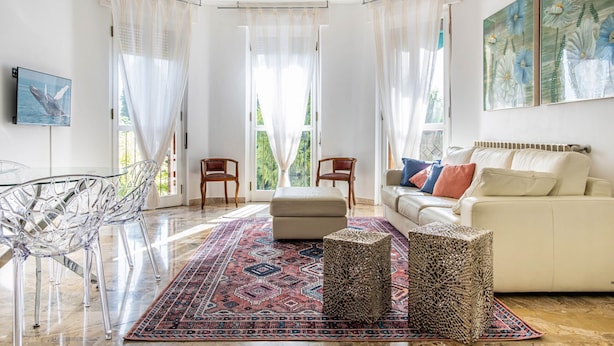 A spacious living room with a beige sofa, two wooden chairs, a glass table, and a patterned rug. - Relais Vittoriale (Gardone Riviera)