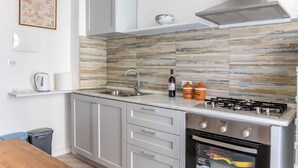 Eine moderne Küche mit Gasherd, Spüle aus Edelstahl und Mikrowelle. Auf der Arbeitsfläche steht eine Weinflasche und zwei orangefarbene Behälter.