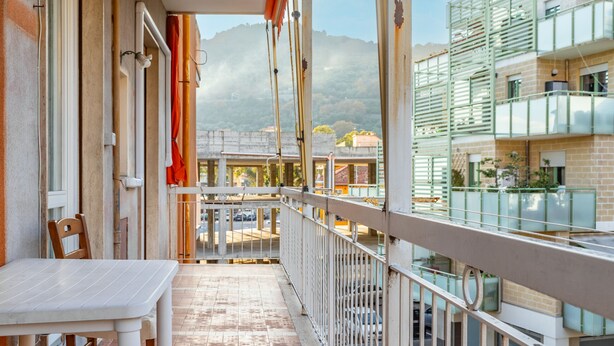 Ein Balkon mit Tisch und Stühlen, der auf eine Straße und umliegende Gebäude mit Bergpanorama blickt.