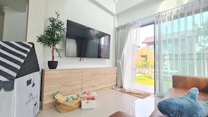 A modern living room with a flat-screen TV, wooden cabinets, a potted plant, and a view of a garden through large windows. - Castello Di Mare (Cha-am)
