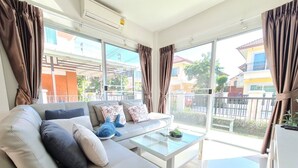 A modern living room with a sofa, cushions, a glass coffee table, and large windows offering a view of the neighborhood. - Castello Di Mare (Cha-am)