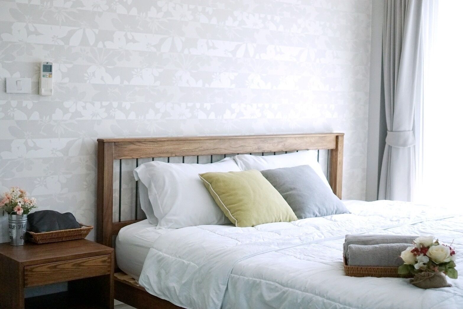 A neatly made bed with a wooden headboard, a bedside table with a basket and a vase of flowers, and a wall with a floral pattern.