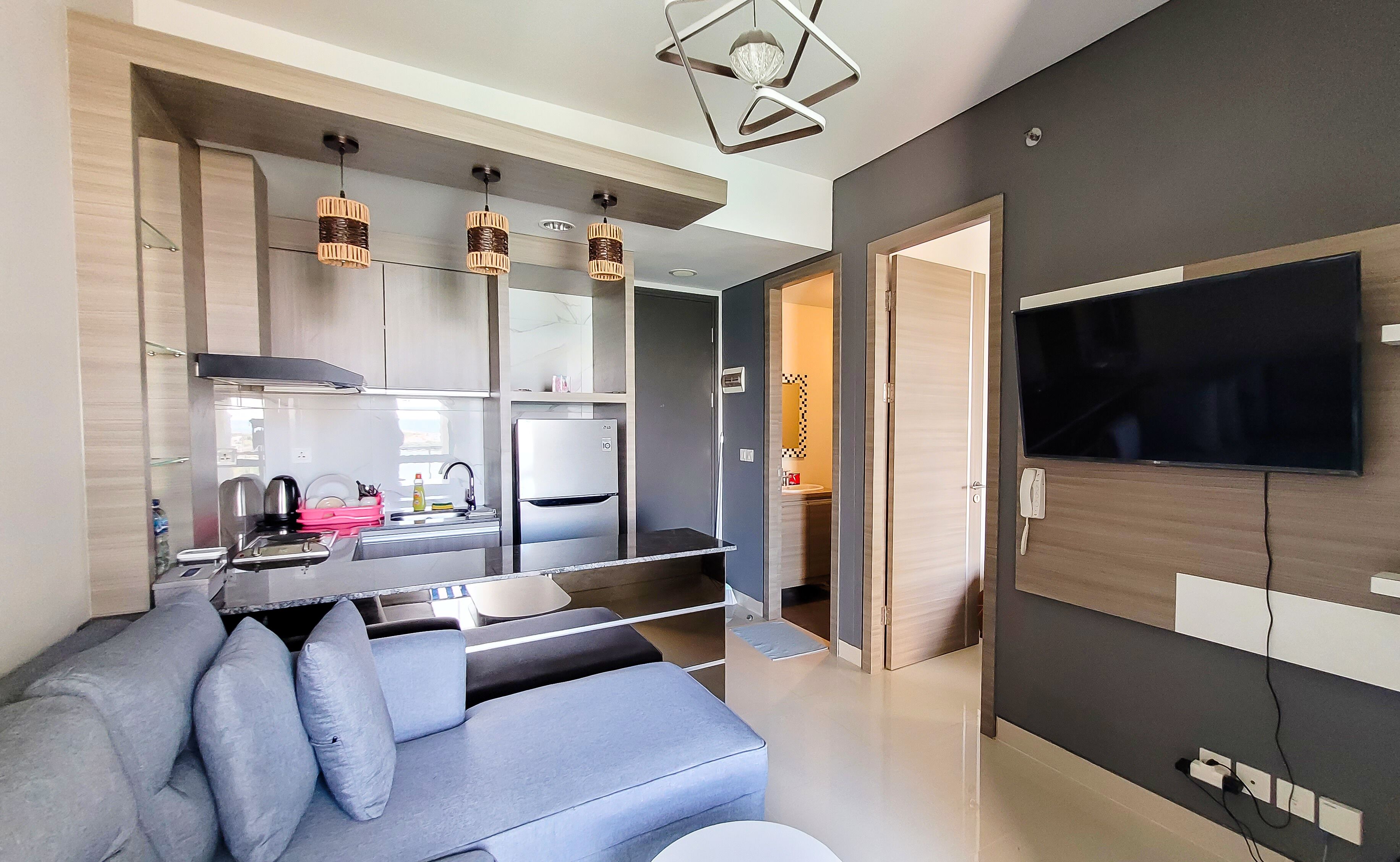 A modern living room with a grey sofa, a flat-screen TV, and a kitchen area with a fridge, stove, and hanging lights.
