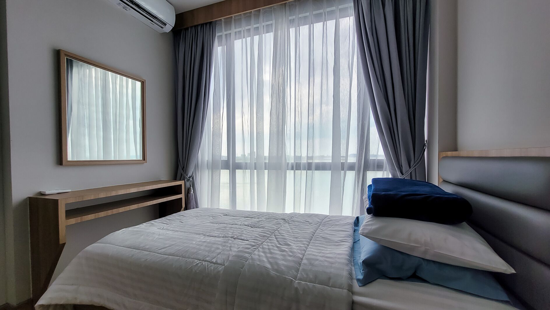 Un dormitorio con cama, mesita de noche, espejo y ventana con cortinas traslúcidas.