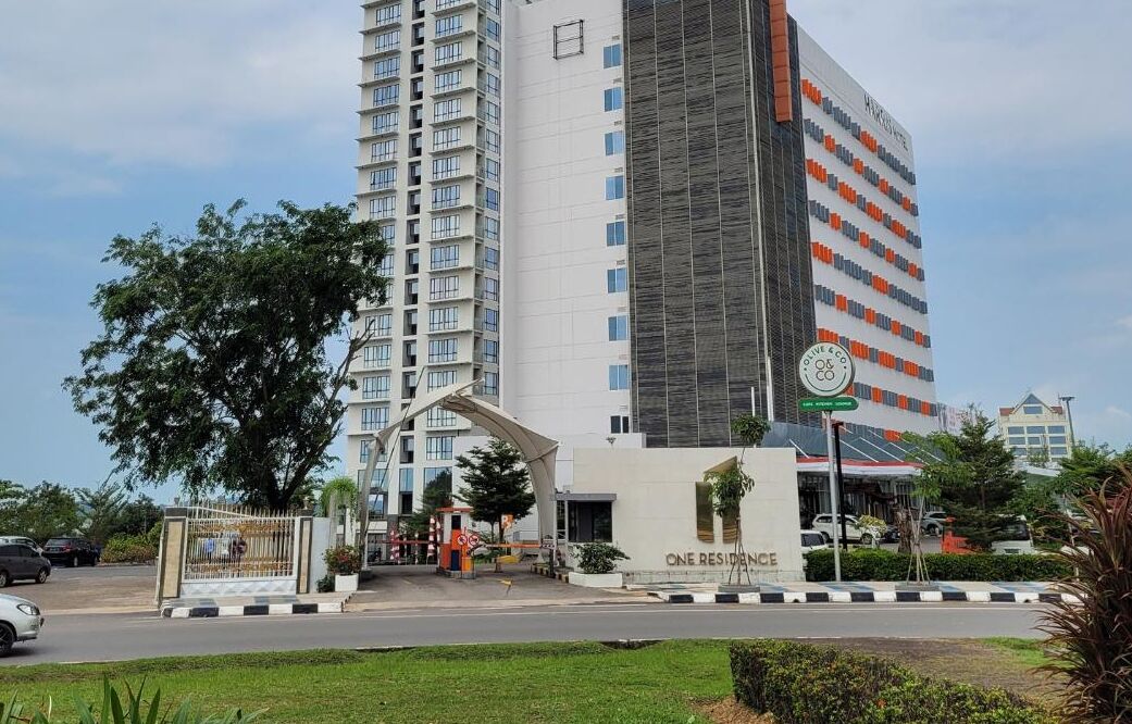 A modern building with a prominent entrance, a sign for 'ONE RESIDENCE', and a parking area with cars.