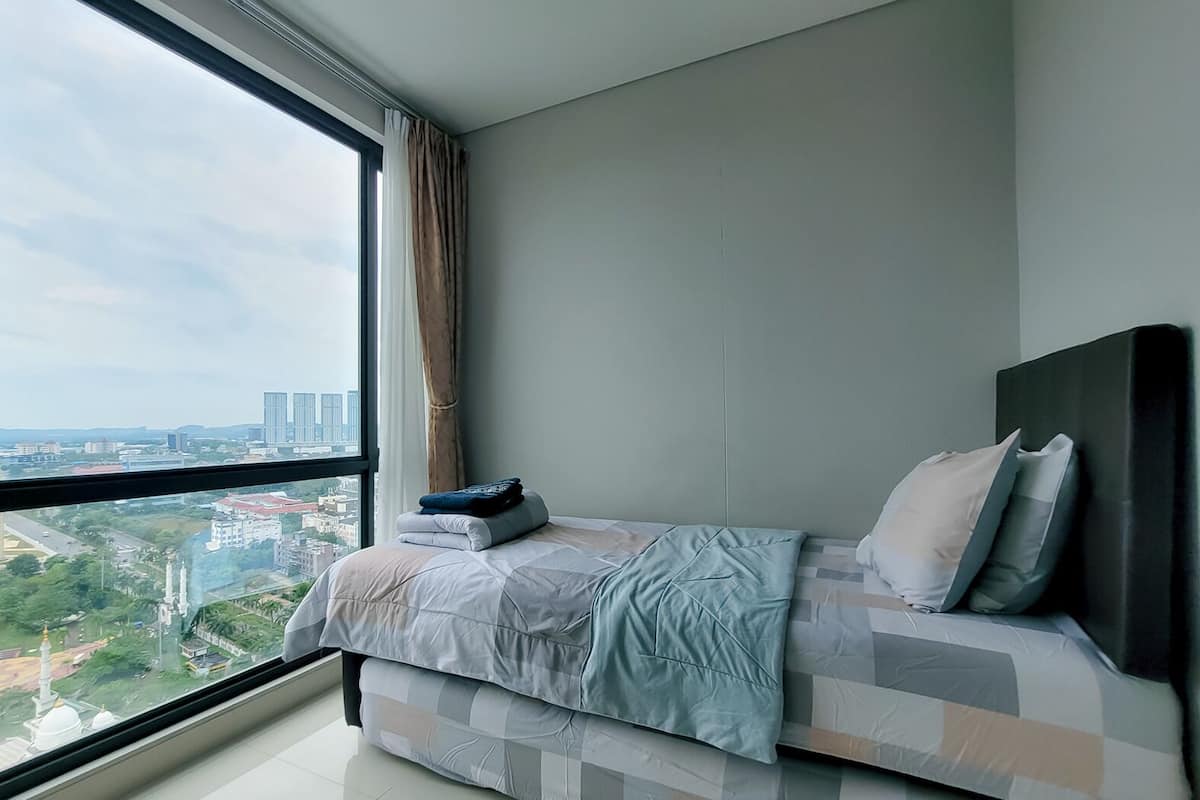 Ein Schlafzimmer mit großem Fenster, einem Bett mit blauer Decke und Blick auf eine Stadtansicht.