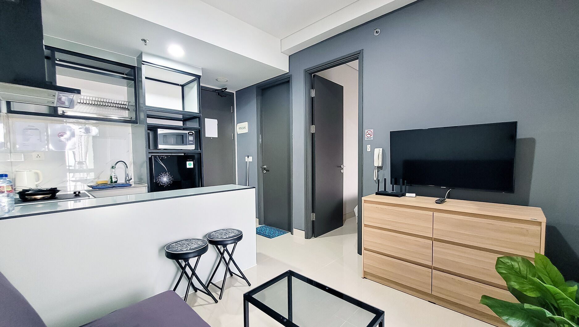 Una cocina moderna con microondas, cafetera y televisor sobre un mueble de madera. Hay dos taburetes y una mesita.