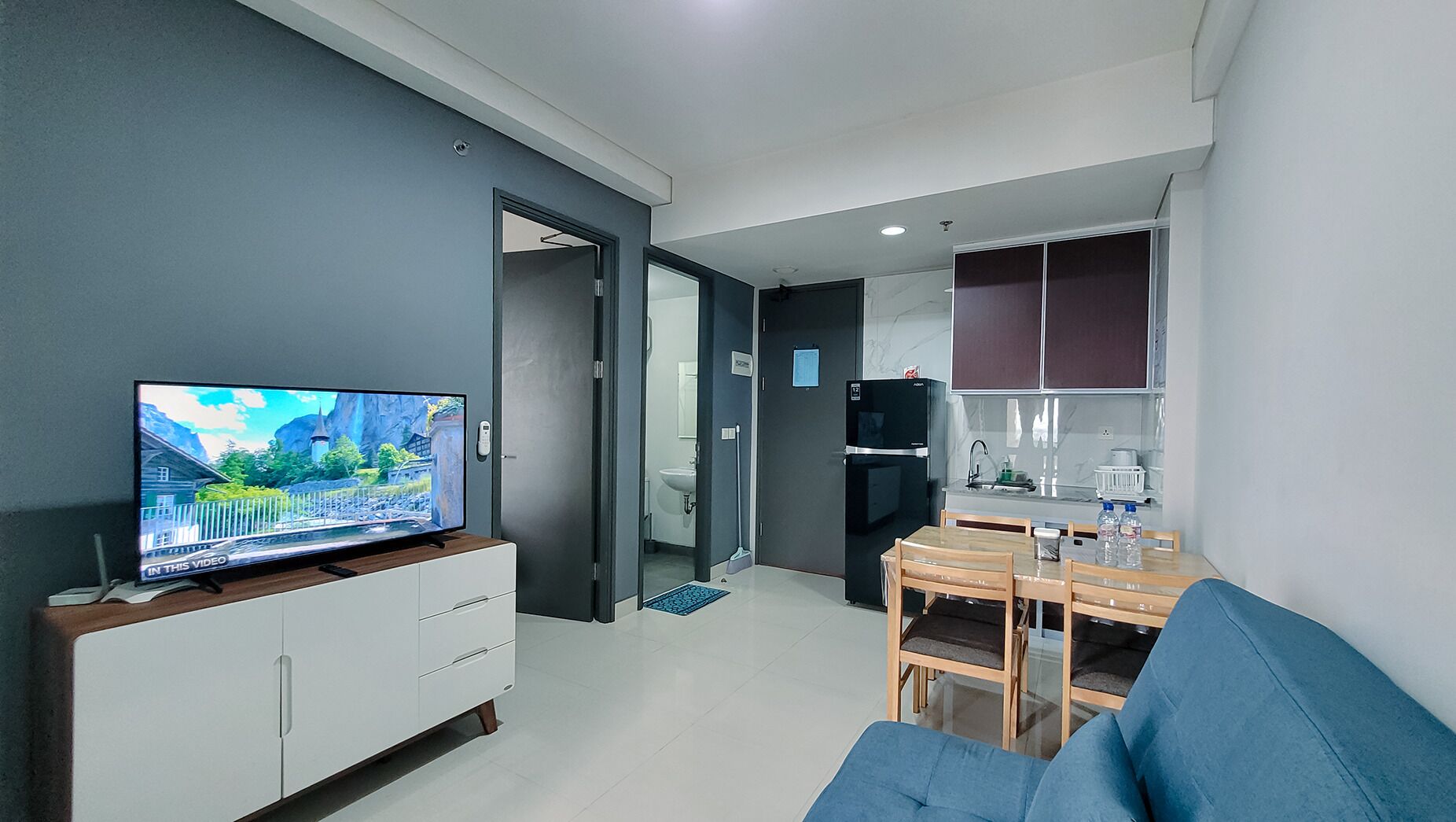 A modern living room with a TV, a sofa, a dining table with chairs, and a kitchen area with cabinets and a refrigerator.