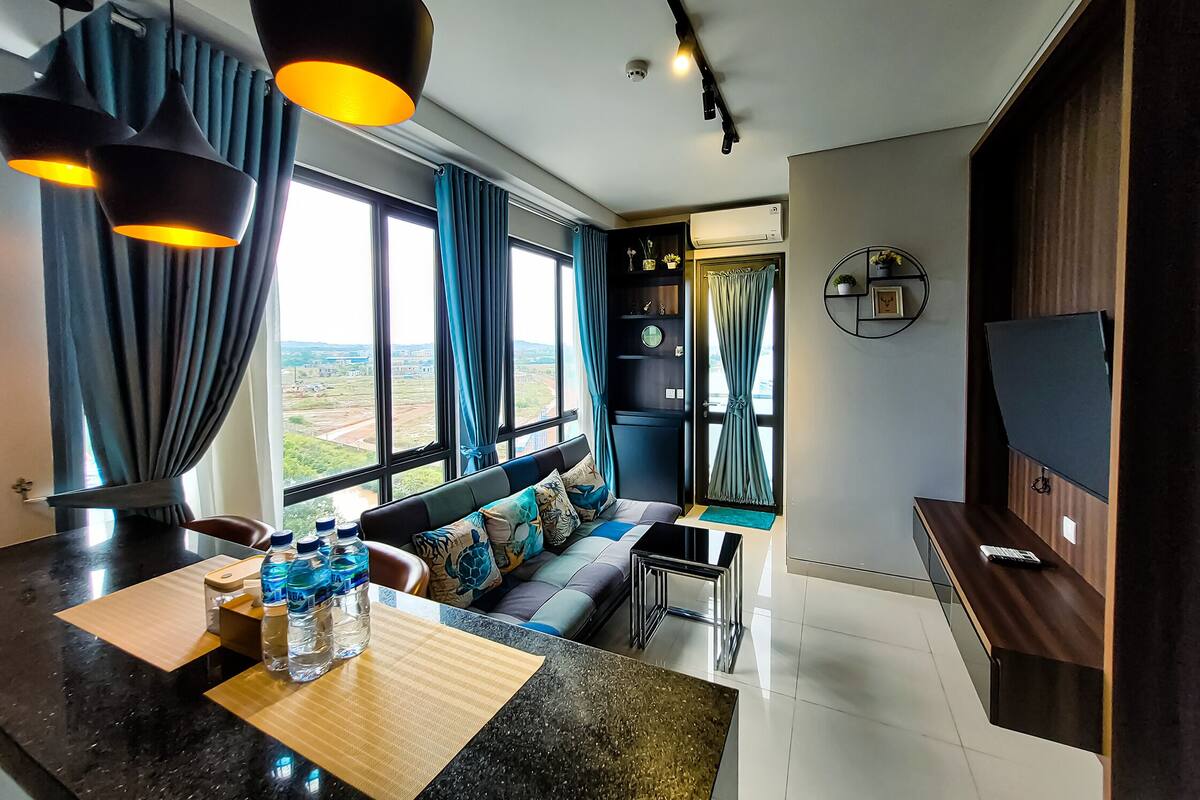 Ein modernes Wohnzimmer mit einer Couch, einem Couchtisch und einem Fernseher. Große Fenster mit Vorhängen, ein Deckenventilator und ein Wasserspender auf dem Tisch.