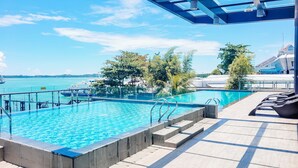 Una piscina con agua azul cristalina, rodeada por una terraza con sillones de descanso, y vistas al mar y a los barcos en el fondo.