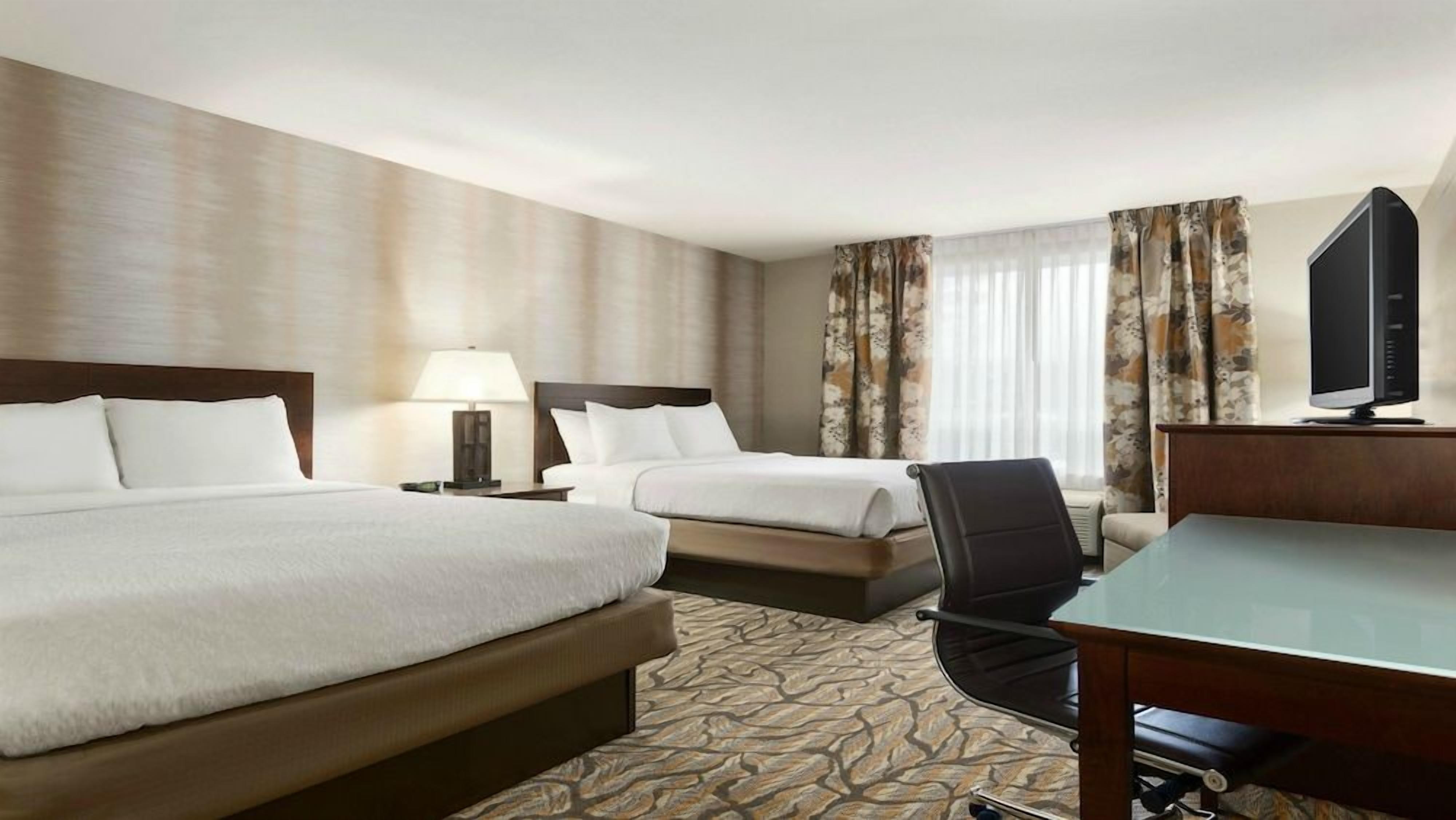 A hotel room with two beds, a desk, a chair, a television, and a window with curtains.