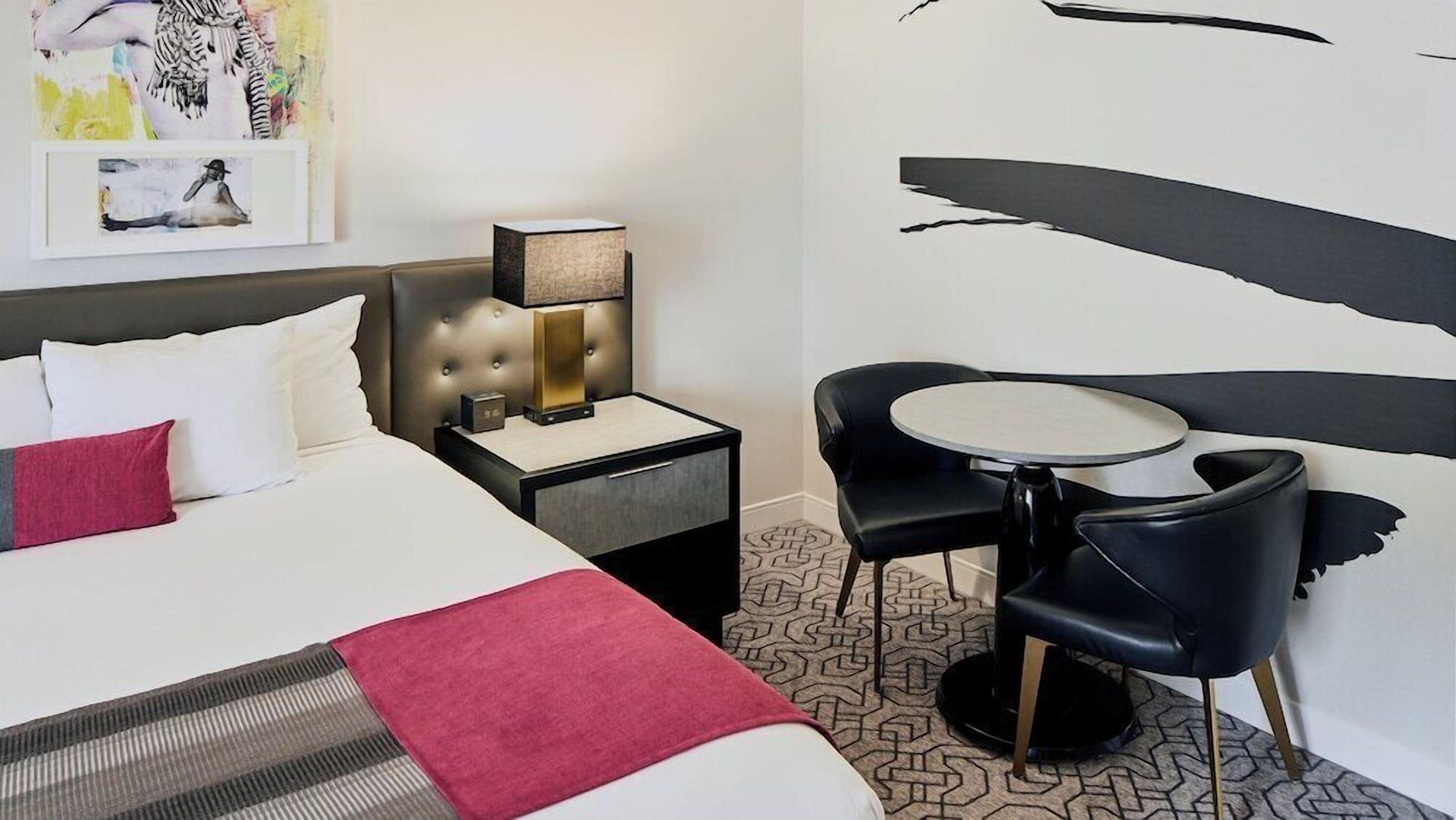 Een hotelkamer met een bed, nachtkastje, lamp en een klein rond tafeltje met twee stoelen.