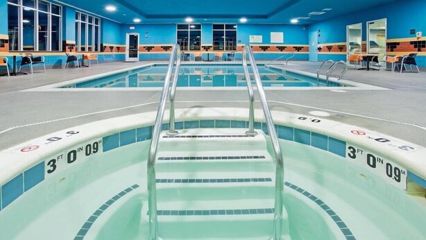 Una piscina con una escalera central, rodeada de sillas y un muro de azulejos.