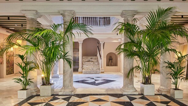 A spacious lobby with a geometric tile floor, marble columns, and potted plants. - Villa de Andas (Cádiz)