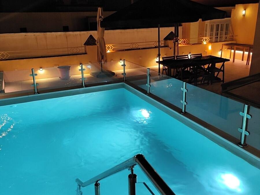 A swimming pool with a glass barrier, a table and chairs, and ambient lighting.
