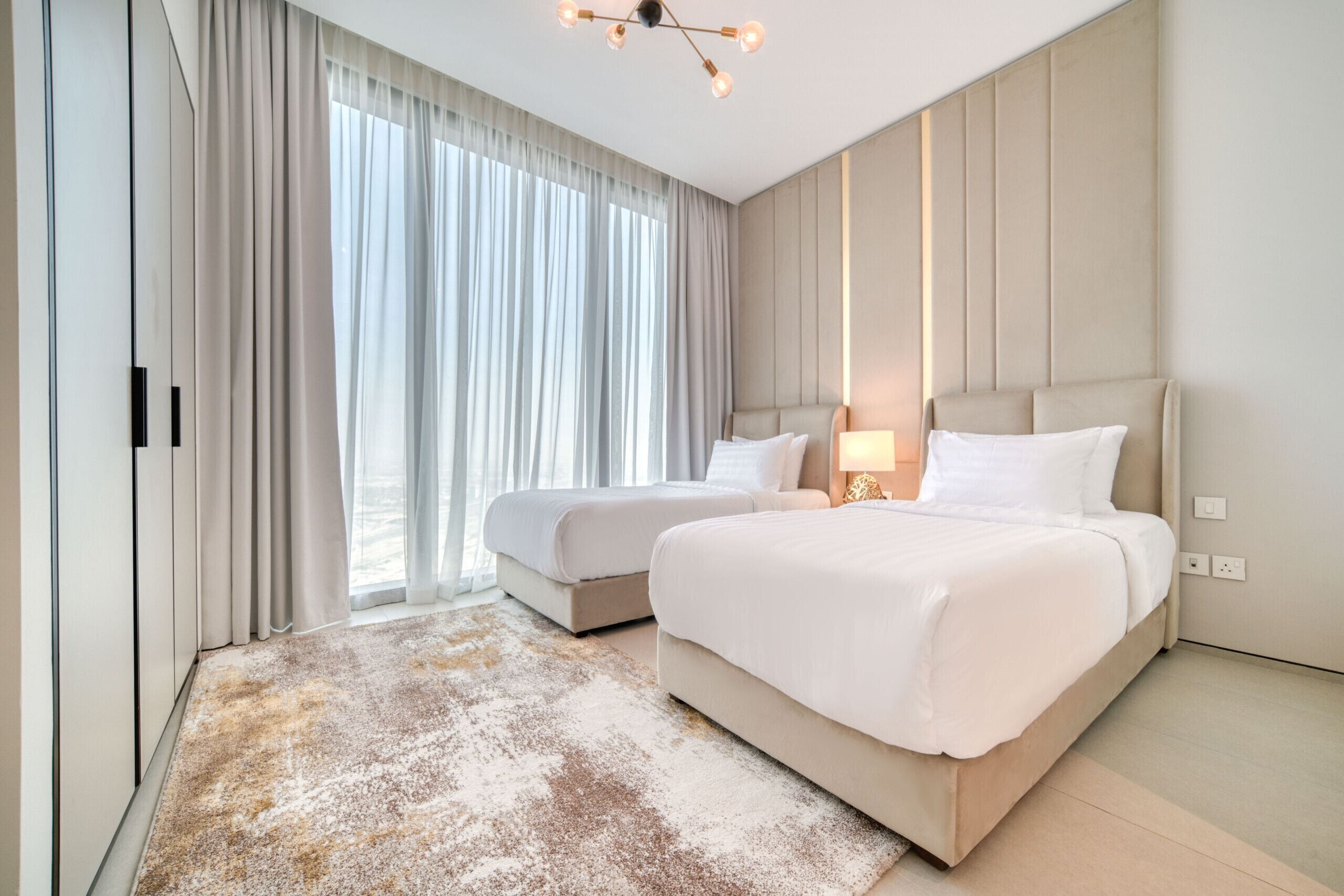 A modern hotel room with two beds, a large window with sheer curtains, a light-colored rug, and a ceiling light fixture.