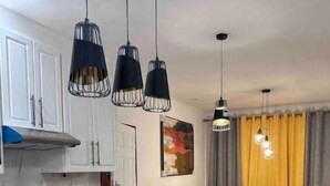 Una cocina moderna con encimera negra, armarios blancos y luces colgantes. Hay un comedor con un sofá y cortinas amarillas.