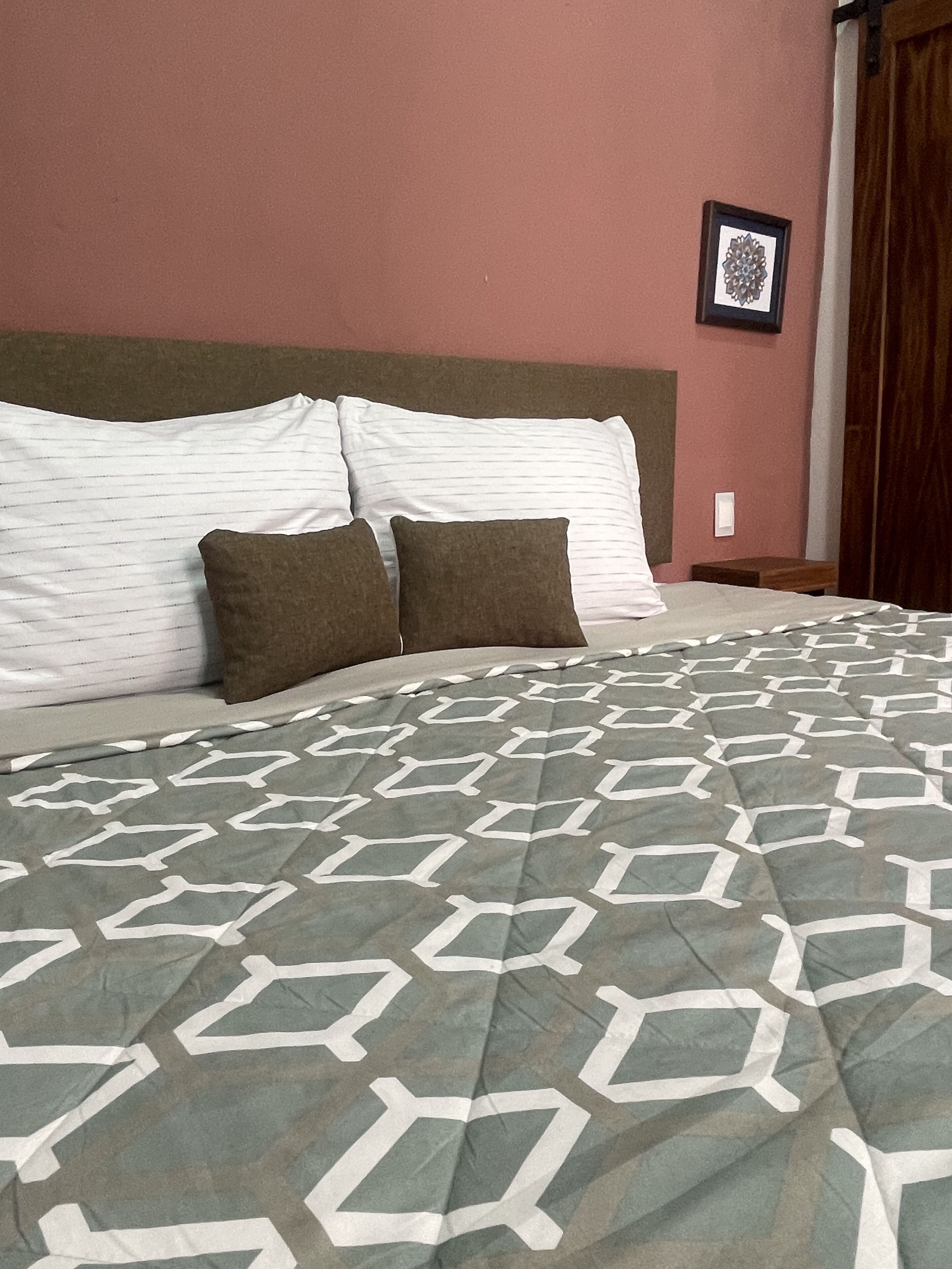 A neatly made bed with a geometric patterned bedspread, two pillows, and a headboard against a pink wall.