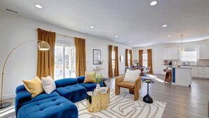 A modern living room with a blue sofa, a round coffee table, and a kitchen area with white cabinets and a central island. - Newly Built Bloomington Oasis: 4bd/2.5br & Backyard, 10mins to IUB & Downtown (Bloomington)