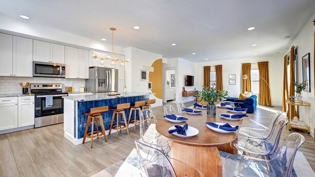 A modern kitchen with white cabinets, a central island with bar stools, and a dining area with a wooden table and clear chairs. - Newly Built Bloomington Oasis: 4bd/2.5br & Backyard, 10mins to IUB & Downtown (Bloomington)