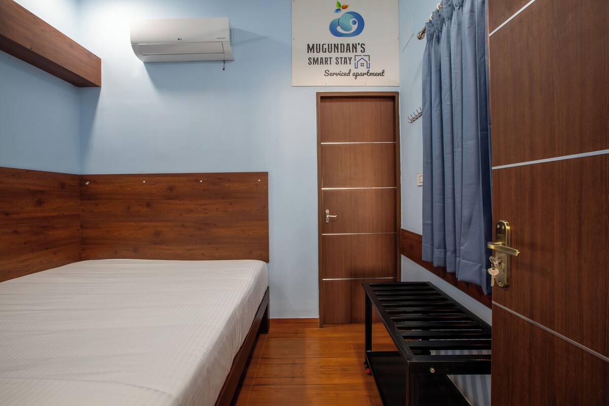 A bedroom with a wooden headboard, a bed, a small bench, a door, and an air conditioning unit.