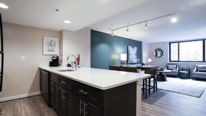 A modern kitchen with dark cabinetry, a white countertop, and a dining area with a table and chairs. - Sojourn | Courtland Towers (Arlington)