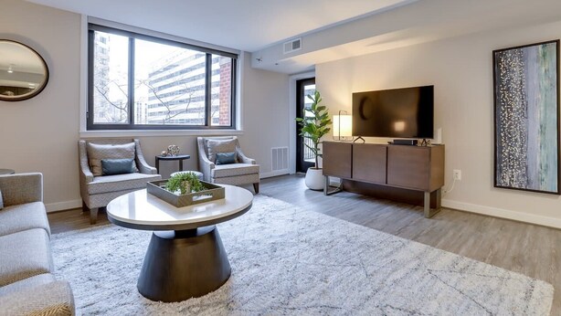 A modern living room with a large window, a round coffee table, a sofa, and a TV on a cabinet. - Sojourn | Courtland Towers (Arlington)